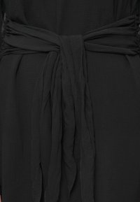 Μαύρο υφασμάτινο υλικό με υφή δεμένο σε χαλαρό κόμπο στη μέση, με μακριές, ρέουσες άκρες να κρέμονται στη μέση.