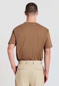 Uomo con capelli corti che indossa una camicia a maniche corte marrone infilata in pantaloni beige, in piedi con una mano in tasca, di spalle rispetto alla fotocamera.