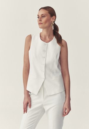 Kvinde med brunt hår i en lav hestehale iført store guldringe, en hvid, ærmeløs vest med knapper og hvide bukser mod en ensfarvet baggrund.