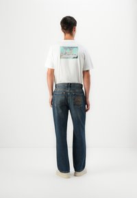 Camiseta blanca con un gráfico colorido de Timberland en la parte posterior, combinada con unos jeans de mezclilla azules que tienen un logo en el bolsillo trasero.