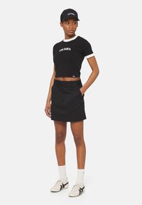 Svart kortärmad t-shirt med vit kant och logotyp, svart kjol med fickor, vita strumpor och bruna sneakers med svarta detaljer. Svart keps.