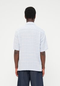 Άνδρας με κοντομάνικο πουκάμισο γαλάζιο και λευκό καρό και σκούρο μπλε παντελόνι, που στέκεται με την πλάτη προς την κάμερα.