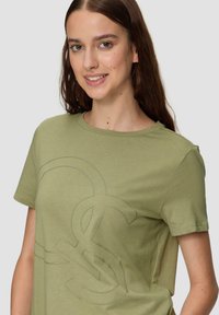 Olijfgroene katoen t-shirt met een groot, reliëflogo in een iets donkerder groen. Ronde halslijn en korte mouwen. Gladde textuur.