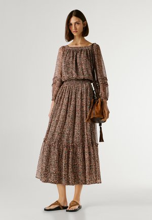 Donna che indossa un abito floreale a maniche lunghe e lunghezza fino alla caviglia con vita smockata e porta una borsa secchiello in camoscio marrone, abbinata a sandali marroni.