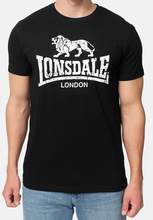 Mann trägt ein schwarzes T-Shirt mit kurzen Ärmeln mit weißem Lonsdale London-Logo und Löwengrafik, kombiniert mit hellblauen Jeans.