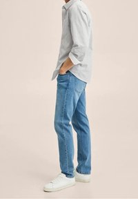 Licht gestreept overhemd met knopen in blauw en wit, gecombineerd met relaxed-fit blauwe spijkerbroek en witte sneakers, weergegeven tegen een neutrale achtergrond.