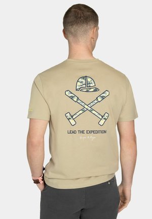 Hombre que lleva una camiseta beige con gráfico de bates de béisbol camuflados cruzados y gorra, texto "Lead the Expedition" en la espalda, de pie con las manos en los bolsillos.