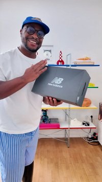Scatola nera delle scarpe New Balance con logo bianco, tenuta in mano. Presenta una forma rettangolare e un coperchio piatto con maniglie laterali sagomate.