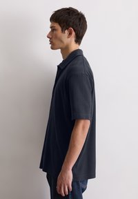 Donkerblauwe geribbelde polo met korte mouwen, knoopsluiting en een relaxte pasvorm, gedragen met donkere jeans, getoond in profielweergave.