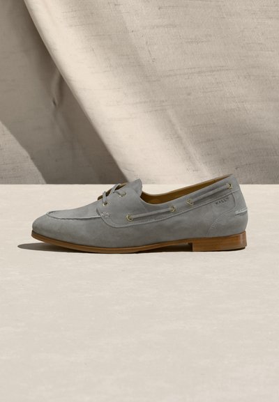 Γκρι μοκασίνι από σουέντ με λεπτομέρεια κορδονιού, ξύλινη σόλα και χαμηλό τακούνι τοποθετημένο σε ανοιχτή επιφάνεια με φόντο από υφασμένο ύφασμα.