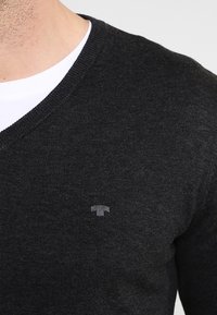 Pull à col en V gris foncé en tissu tricoté texturé, arborant un petit logo sur la poitrine et porté par-dessus un T-shirt blanc.