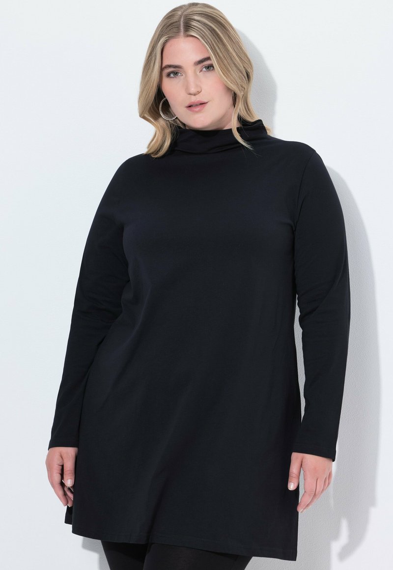Eine Frau mit blonden Haaren trägt ein schwarzes, langärmliges Rollkragenkleid und steht vor einer weißen Wand.