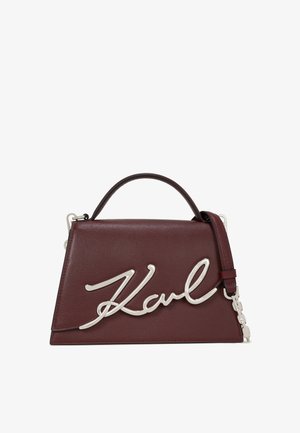 Bolsa de mão em couro bordeaux com design angular, apresentando um logótipo em metal prateado "Karl" e uma alça de ombro destacável. Acabamento texturizado.