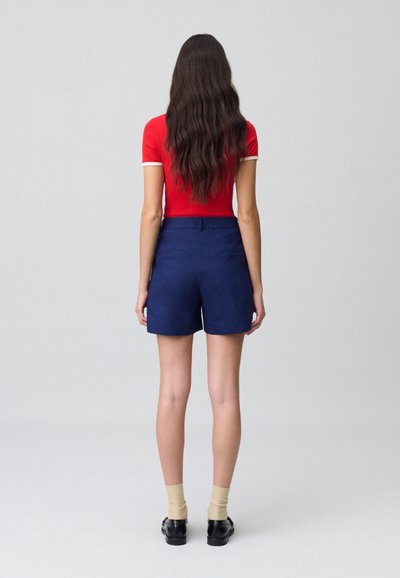 Top corto de mangas cortas de rayas rojas con ribete blanco, combinado con pantalones cortos de talle alto en azul marino oscuro. La modelo lleva calcetines beige claros y zapatos negros.