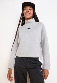 Felpa grigia corta con colletto alto, caratterizzata da un piccolo logo nero. Indossata con pantaloni neri e un cappellino nero, presenta una vestibilità rilassata.