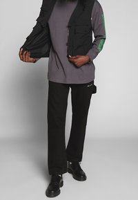 Chaleco negro de múltiples bolsillos sobre una camisa de manga larga gris, combinado con pantalones negros y zapatos negros, que muestra un diseño funcional y un aspecto casual.