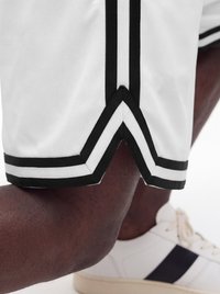 Close-up of a person’s knee bent wearing white shorts with black trim and white sneakers with black stripes on a white background.
