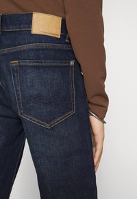 Tmavě modré džíny z denimu s koženou značkovou nášivkou na pasu, s oranžovým prošíváním a jednou zadní kapsou se zakřiveným designem.