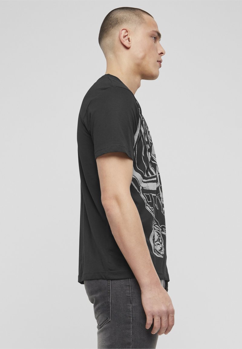 Junger Mann mit kurzen Haaren, der ein schwarzes T-Shirt mit Grafik und dunkle Jeans trägt, steht im Profil vor einem einfarbigen grauen Hintergrund.