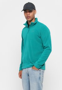 Moški oblečen v teal polovični zip flis, črno kapo in svetlo modre kavbojke, stoji s pripetim rokom v žepu ob enotni ozadju.