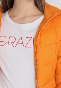 Orange vadderad jacka med dragkedja och en quiltad design. Ovanpå en vit T-shirt med "GRAZI" i fetstil röda bokstäver.