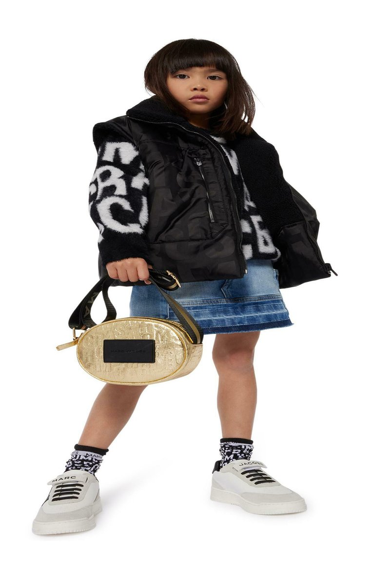 Zwarte pufferjas over een wit en zwart geblokt trui, spijkerrok en grove witte sneakers. Houdt een gouden ovale tas vast met een zwarte accent.