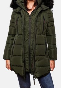 Femme portant une veste d'hiver matelassée vert olive avec plusieurs poches zippées, une capuche bordée de fourrure noire, et un jean bleu.