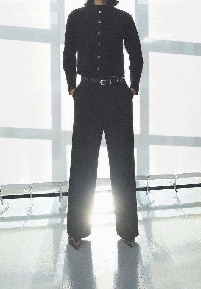 Personne portant une veste noire boutonnée et un pantalon à jambes larges, les mains dans les poches, debout devant une fenêtre lumineuse avec des ombres.