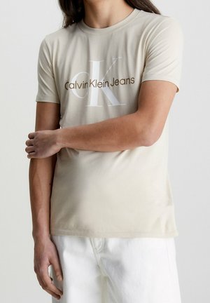 T-shirt en coton beige à manches courtes, avec un grand logo 'CK' blanc et le texte marron "Calvin Klein Jeans" à l'avant.