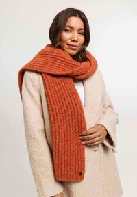 Bufanda acanalada color óxido, hecha de un tejido suave y texturizado, colgada alrededor del cuello, combinada con un abrigo color crema. Presenta una etiqueta de acento negra.