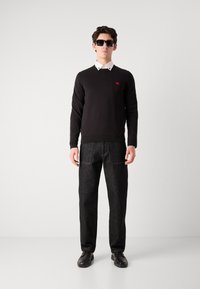 Pulover negru din tricot cu guler rotund, cămașă albă cu guler dedesubt, asortat cu pantaloni de blugi închis la culoare și pantofi negri. Ochelari de soare pătrați purtați.