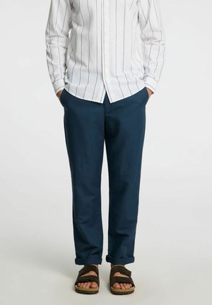 Pantaloni azzurri chiari da uomo in cotone, con vestibilità rilassata, orlo arrotolato, indossati con una camicia a righe bianche e sandali.