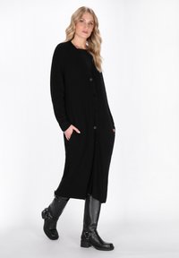 Cardigan lungo nero a costine con chiusura a bottoni e tasche, abbinato a stivali neri al ginocchio con accenti di hardware decorativo.