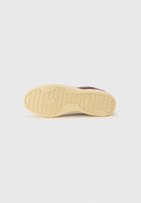 Sole of a sneaker with beige rubber tread featuring geometric patterns, visible part of a burgundy upper, on a white background.