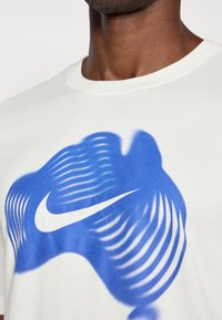 Κοντινό πλάνο ενός ατόμου που φοράει ένα λευκό πουκάμισο με ένα κυματιστό μπλε γραφικό και το λευκό λογότυπο Nike στο στήθος.