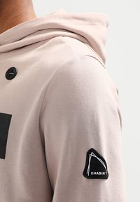 Felpa beige con cappuccio e finitura testurizzata, dotata di un bottone nero e una toppa a forma di triangolo nera sulla manica sinistra.