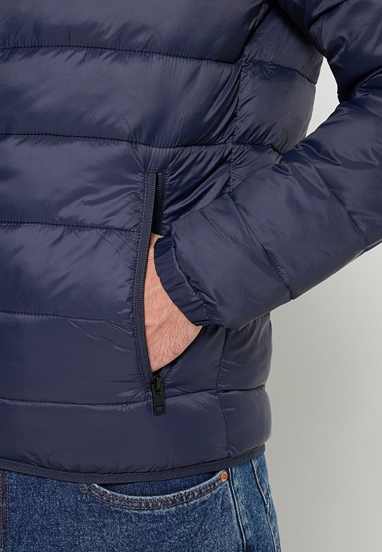 Granatowa kurtka puchowa z pikowanym wzorem, poziomymi sekcjami, zamykanymi na zamek bocznymi kieszeniami, elastycznymi mankietami i prostym dołem. Lekka, gładka w dotyku.