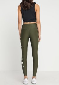 Leggings verde oliva de tiro alto con el texto blanco "SOLO HAZLO" en el lado izquierdo, combinados con una camiseta de tirantes ajustada negra y zapatillas blancas.