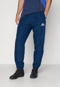 Calças de desporto azul marinho com punhos elásticos e riscas laterais. Feitas de um tecido leve, apresentando o logo da Adidas na perna esquerda.