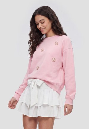Ung kvinde iført en pink sweatshirt med blomsterdetaljer og et hvidt lagdelt skørt, bundet med en sløjfe i taljen, smilende og kiggende ned.