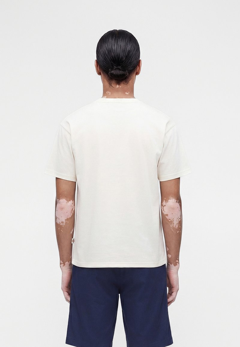 Person mit dunklen Haaren, zu einem Dutt gebunden, trägt ein schlichtes weißes Kurzarmshirt und marineblaue Shorts, steht mit dem Rücken zur Kamera auf weißem Hintergrund.