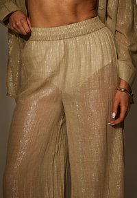 Beige, hoge taille, geplooide broek met een glanzende textuur en metalen draadjes, gecombineerd met een los zittend overhemd met knopen.