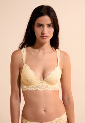 Donna con lunghi capelli scuri che indossa un reggiseno in pizzo giallo chiaro e biancheria intima abbinata, in piedi davanti a uno sfondo beige.