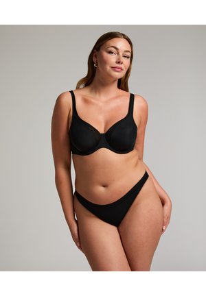 Woman wearing black underwire bra and matching black bikini-style underwear, standing against plain grey background, posing confidently.