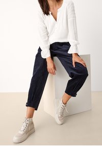 Blusa bianca con maniche lunghe e dettagli smock, abbinata a pantaloni corti navy con texture e sneakers alte beige.