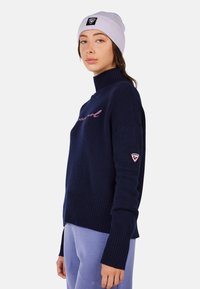 Navy knitted sweater with a high collar, ribbed hem, and shoulder. Features pink embroidered text and a logo patch on the sleeve.