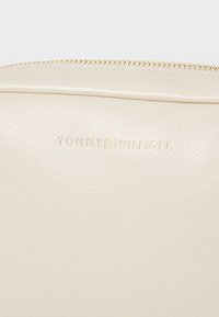Sac en simili cuir beige avec quincaillerie dorée, doté d'une fermeture éclair et du logo « TOMMY HILFIGER » en relief dans une police moderne et épurée.