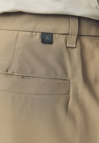 Kerged pruunid püksid siledast materjalist, tagumise taskuga ja musta logo sildiga vöökohtade lähedal.