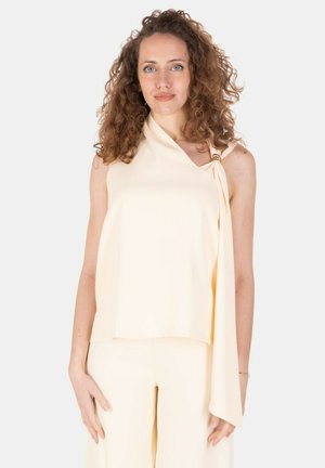 Donna con capelli ricci che indossa un top crema senza maniche con un fiocco sulla spalla e pantaloni crema abbinati, in piedi davanti a uno sfondo semplice.