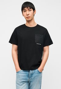 Μαύρο βαμβακερό T-shirt με υφασμένη μαύρη τσέπη που φέρει το λογότυπο «MICHAEL KORS». Κοντά μανίκια και χαλαρή εφαρμογή. Φορεμένο με ανοιχτόχρωμο μπλε τζιν.